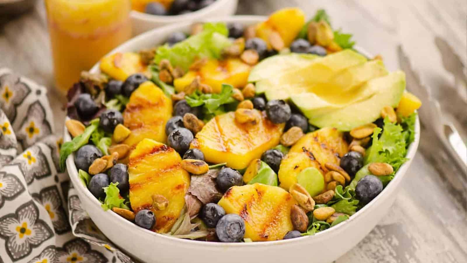 A salad bowl with grilled peaches, blueberries, avocado slices, greens, and seeds on a patterned cloth.
