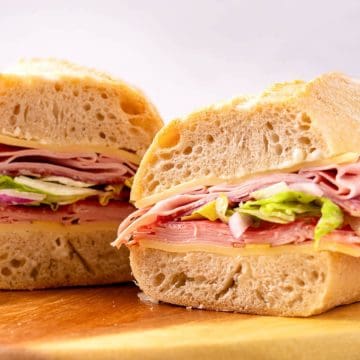 Italian sub sandwich with meats, cheese, and lettuce on thick bread, cut in half and set on a wooden board.