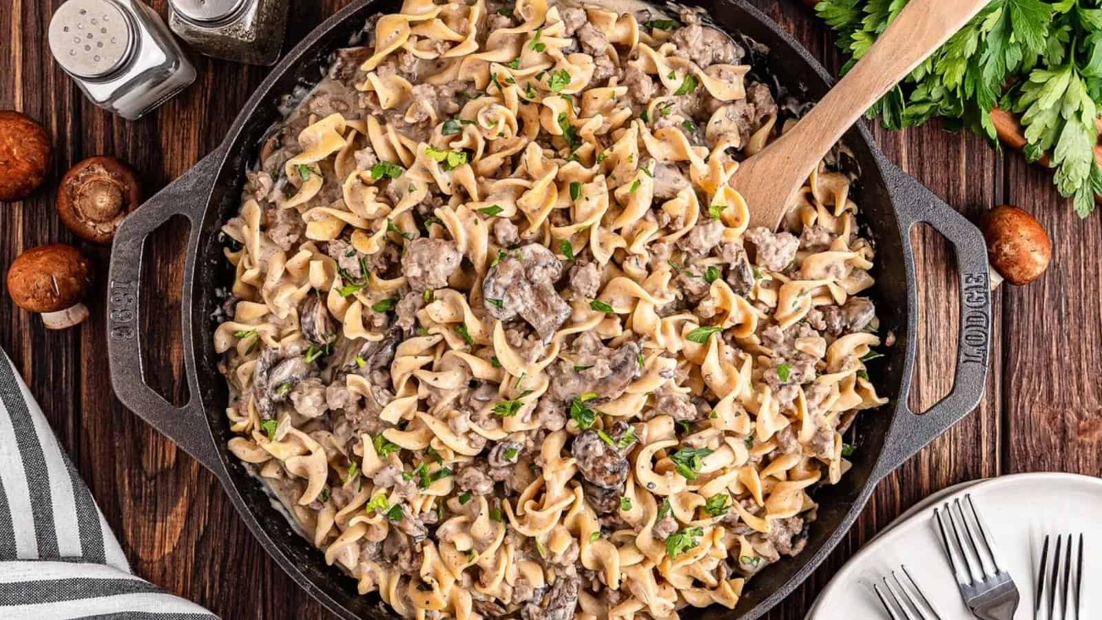 A skillet of creamy beef stroganoff with egg noodles and mushrooms, garnished with parsley and a wooden spoon.