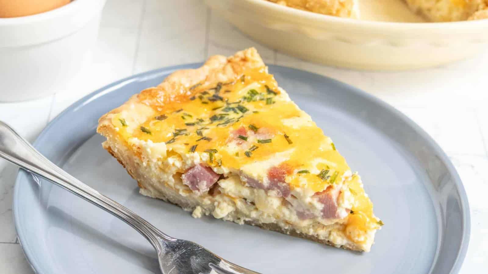 A slice of ham and cheese quiche on a gray plate with a fork, topped with chopped herbs.