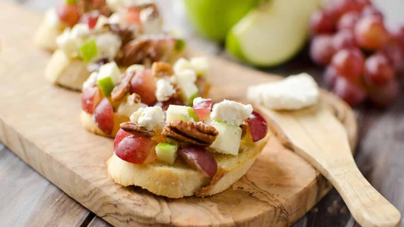 Toasted bread topped with grapes, green apples, pecans, and crumbled cheese on a wooden board.