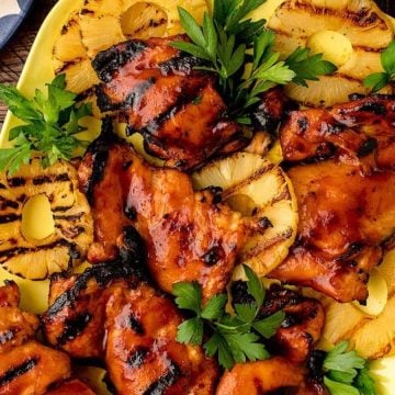 Grilled chicken thighs with barbecue sauce and pineapple rings, garnished with parsley on a yellow platter.