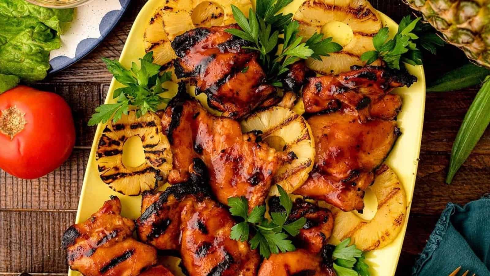 Grilled chicken thighs with barbecue sauce and pineapple rings, garnished with parsley on a yellow platter.