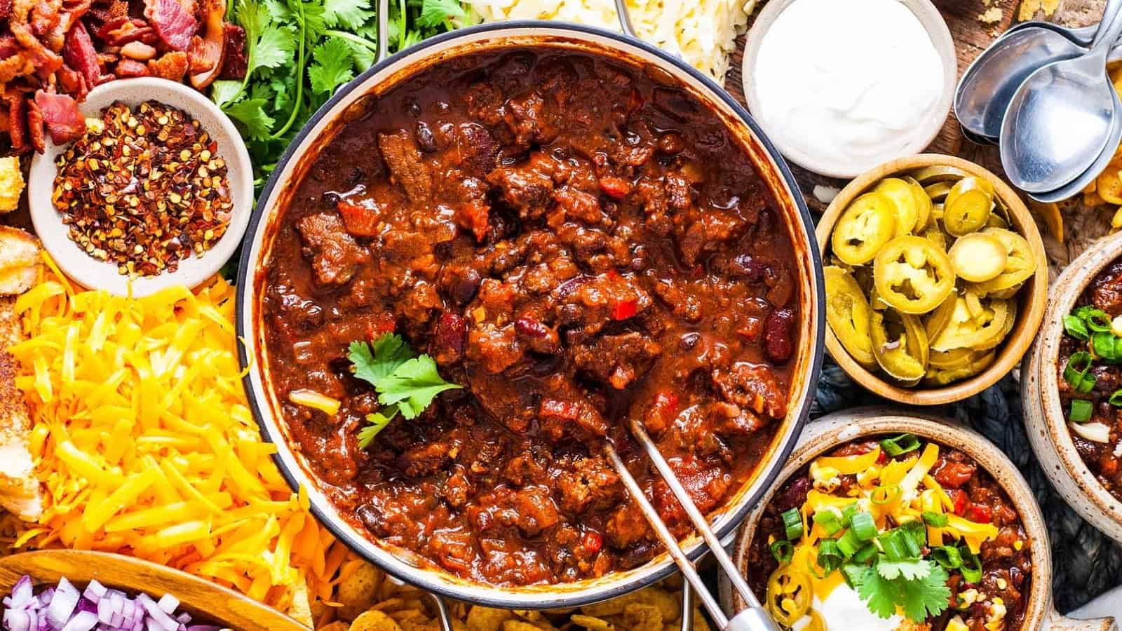 A bowl of chili with toppings like cheese, jalape&ntilde;os, onions, sour cream, and herbs arranged around it.