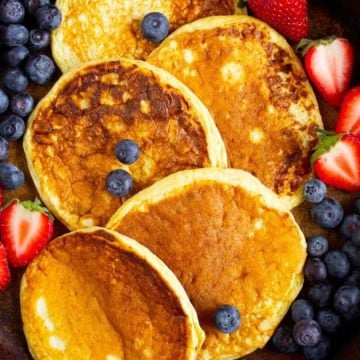 Four pancakes on a plate with fresh blueberries and halved strawberries around them.
