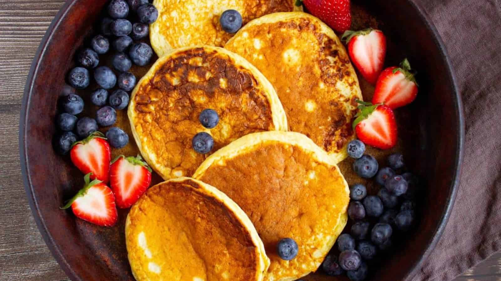 A pan with five high protein cottage cheese pancakes surrounded by blueberries and halved strawberries on a wooden table.