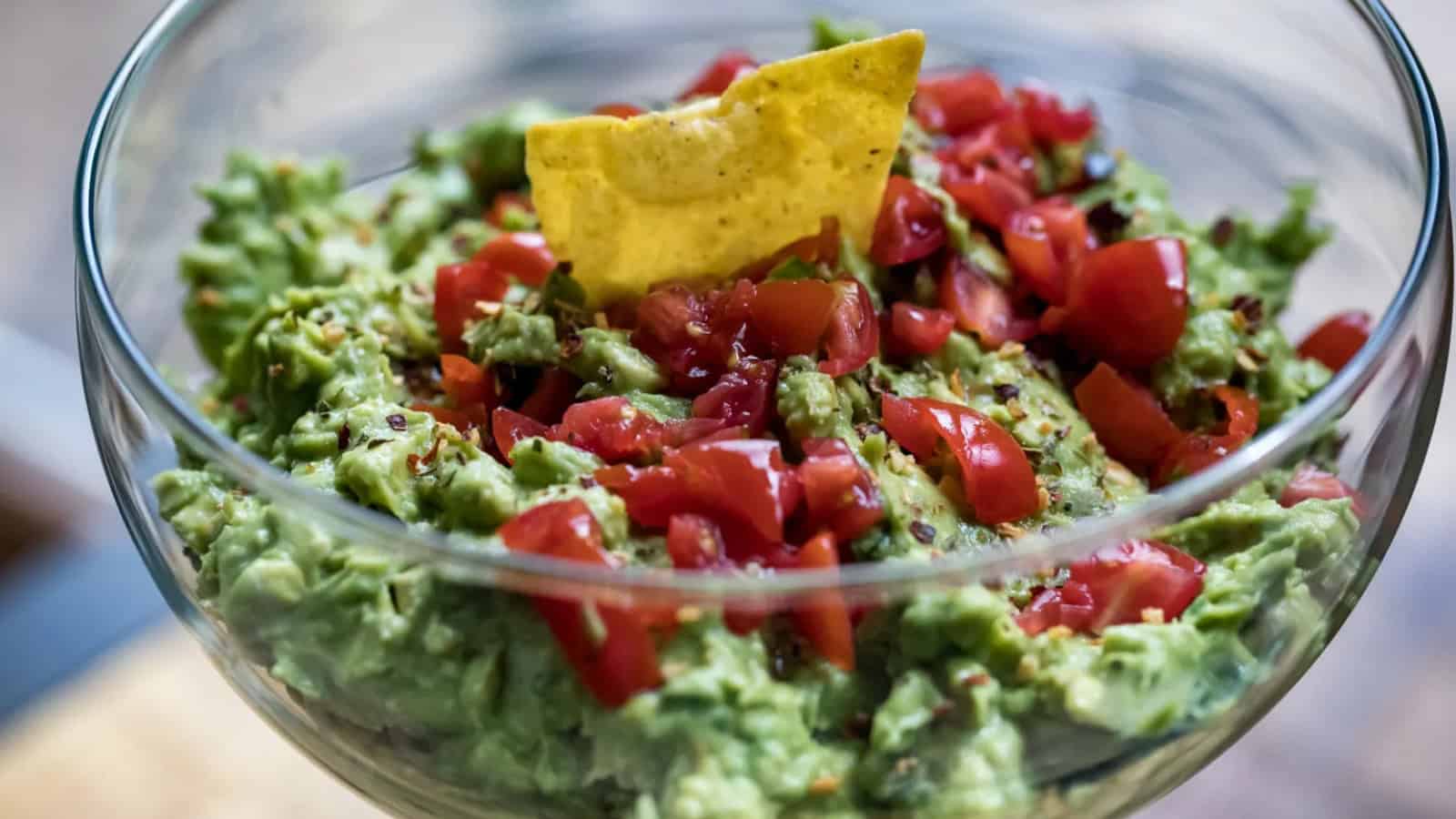Bowl of guacamole topped with diced tomatoes and a tortilla chip standing upright in the center.