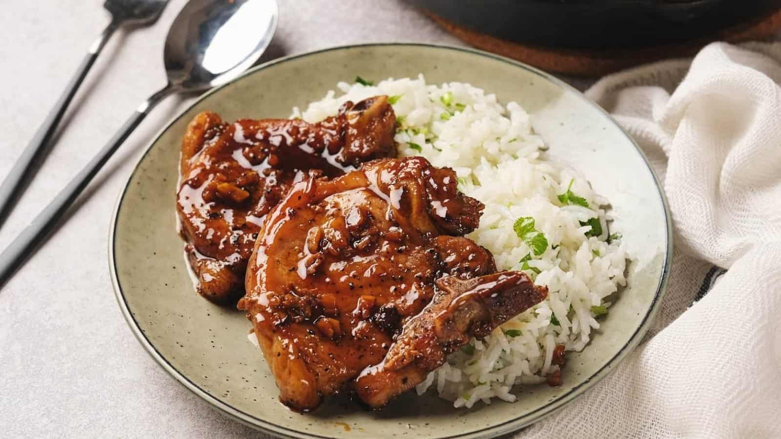 A plate with honey garlic pork chops glazed with sauce, accompanied by a serving of rice garnished with herbs. A fork and spoon are placed beside the plate.