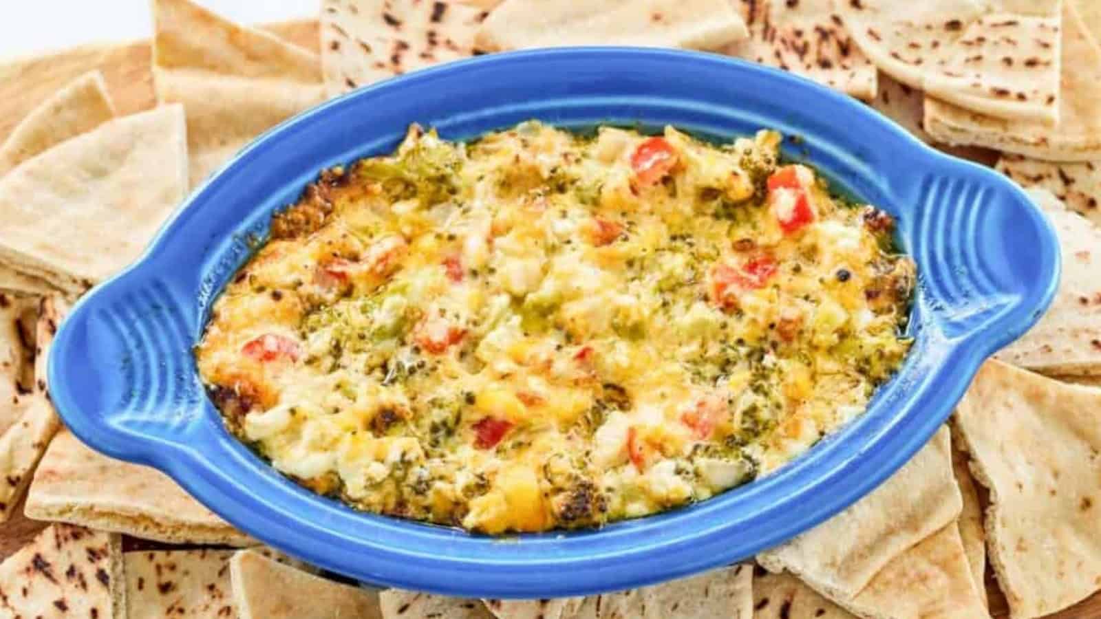 Warm cheesy dip with red and green peppers in a blue dish, surrounded by triangles of pita bread.