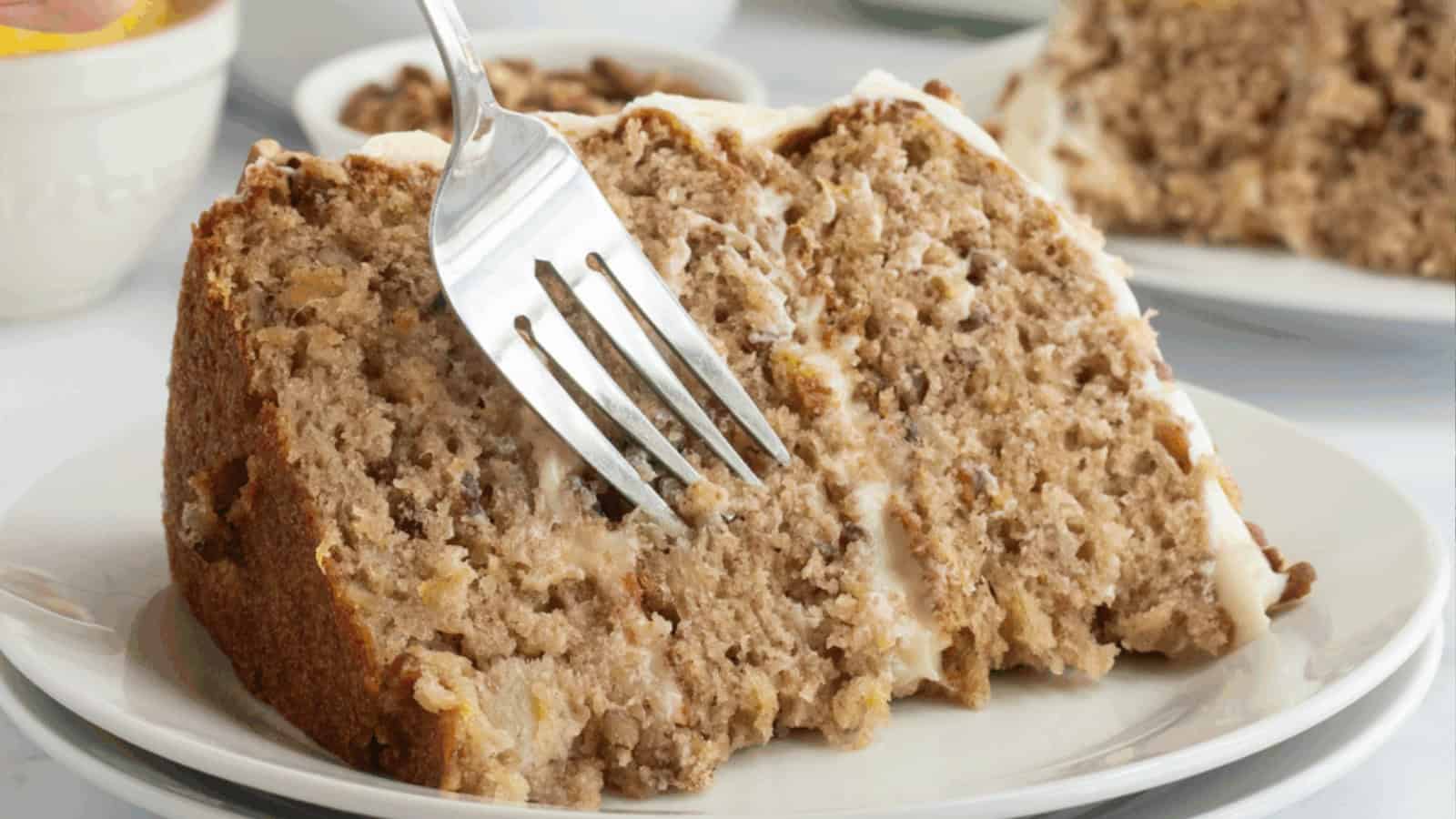A fork cutting into a slice of moist, frosted cake with nuts on a white plate.