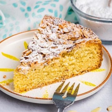 A slice of almond cake topped with powdered sugar on a plate, surrounded by raspberries and sliced almonds.