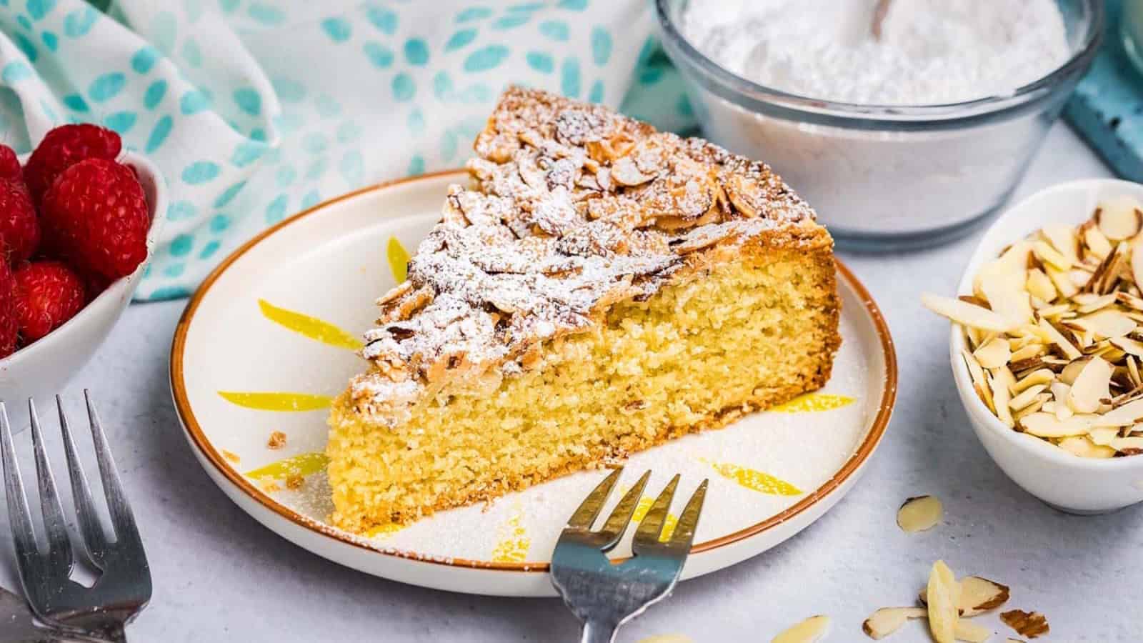 A slice of almond cake topped with powdered sugar on a plate, surrounded by raspberries and sliced almonds.