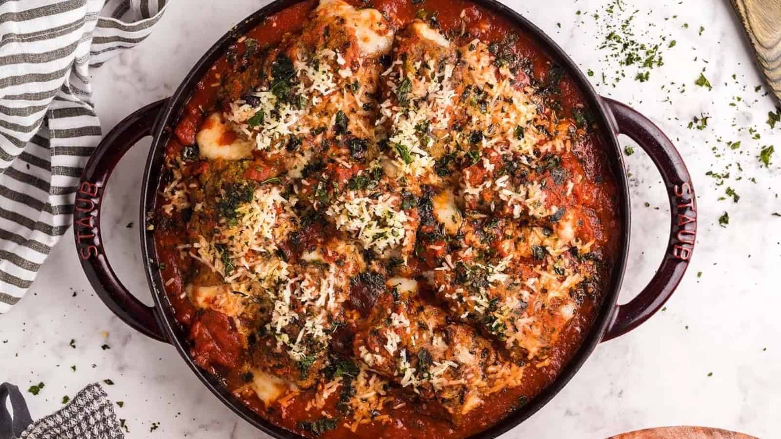 Baked chicken Parmesan in a round red dish, topped with melted cheese, herbs, and tomato sauce.