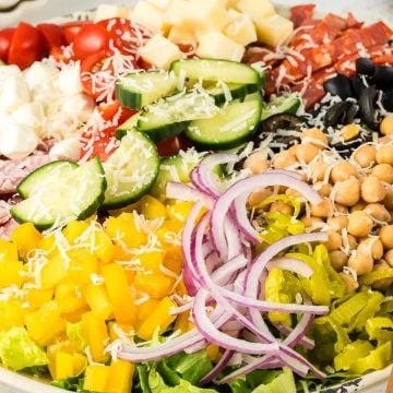 A bowl of chopped salad with lettuce, cucumbers, tomatoes, chickpeas, cheese, meats, olives, and onions.