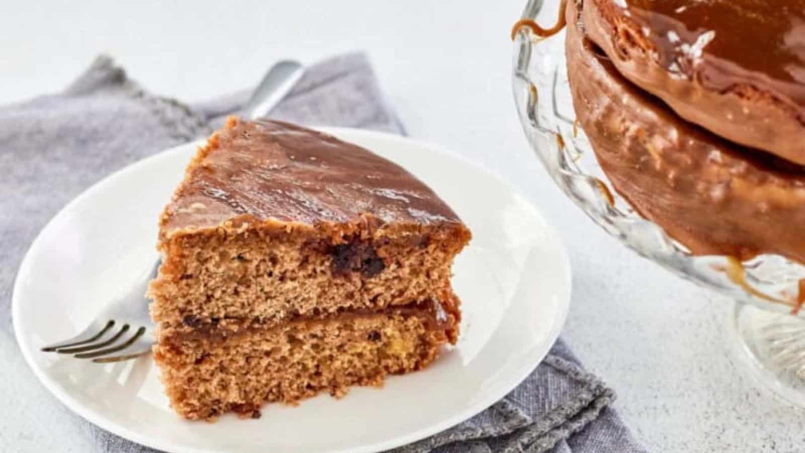 A slice of chocolate-frosted cake on a white plate with a fork, next to the whole cake on a stand.