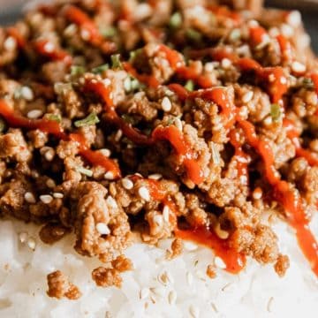 Close-up of white rice topped with seasoned ground meat, sesame seeds, and a drizzle of red sauce.