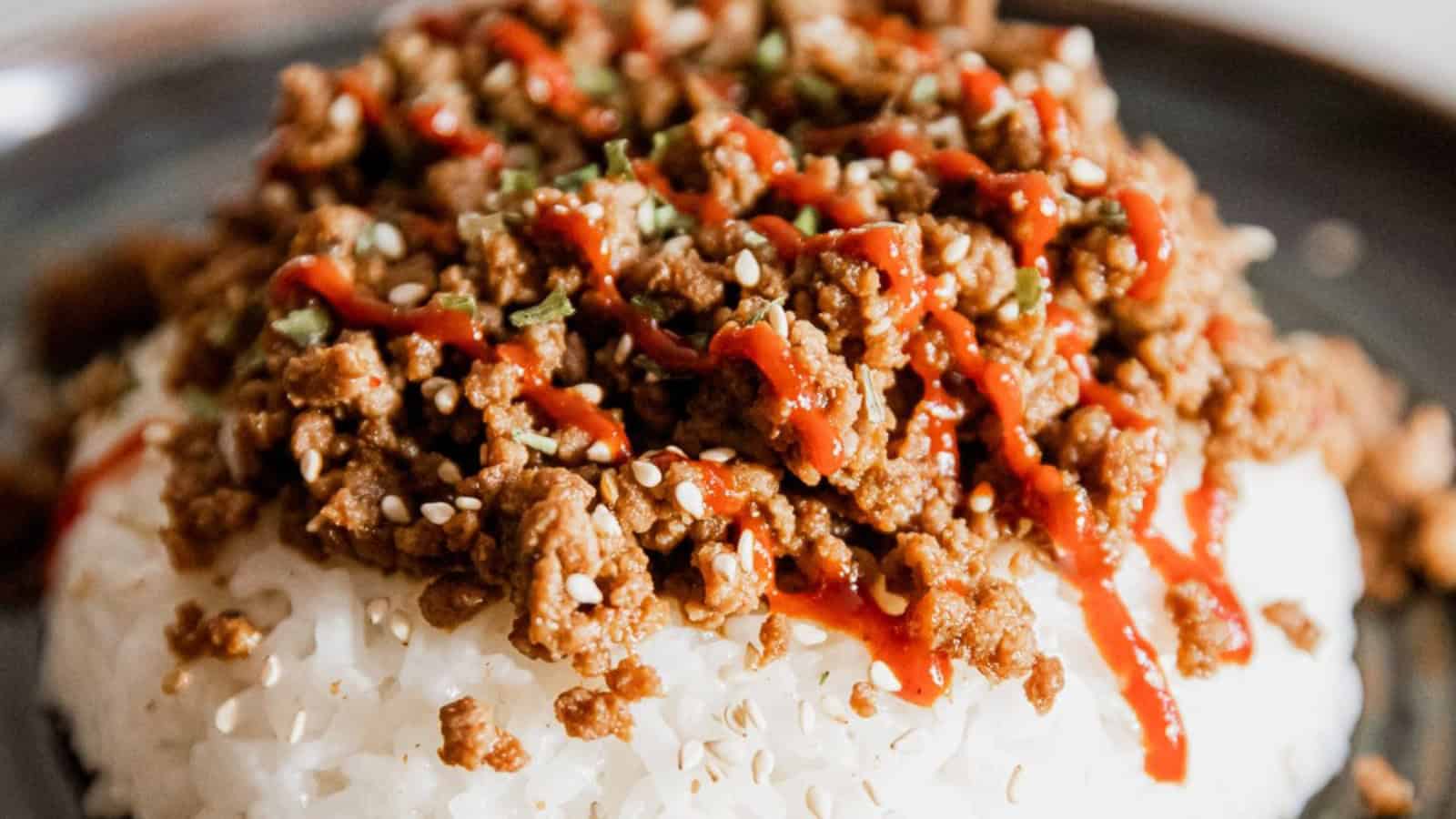 Close-up of white rice topped with seasoned ground meat, sesame seeds, and a drizzle of red sauce.