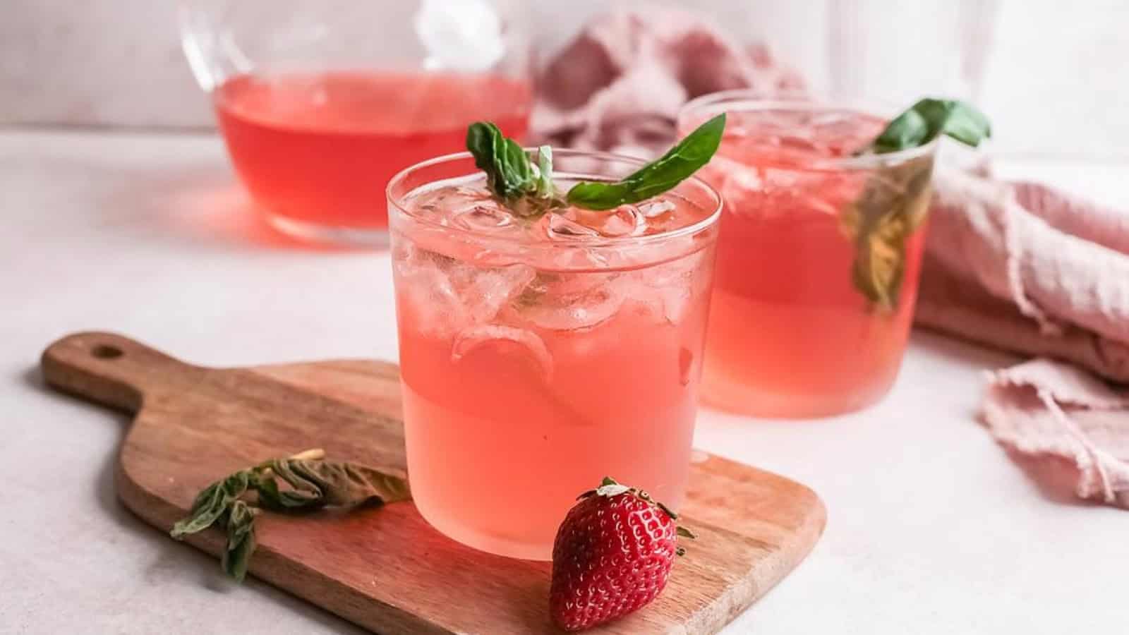 Two glasses of pink iced drink with basil, a strawberry, and a pitcher in the background on a wooden board.