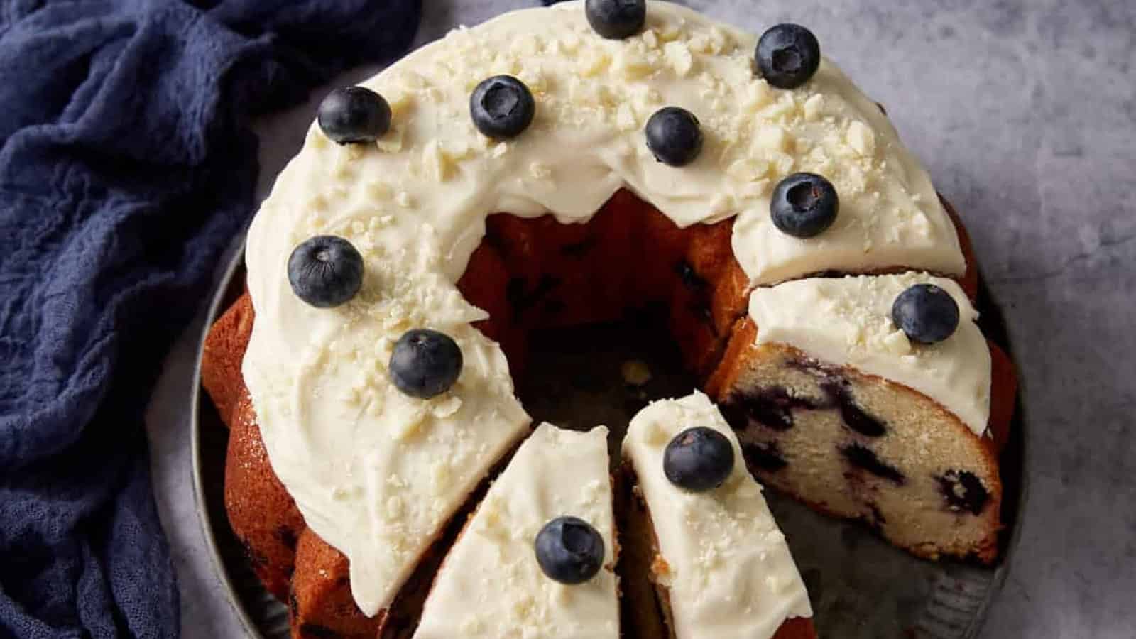 A bundt cake with white icing, topped with blueberries and white chocolate shavings, with one slice cut.