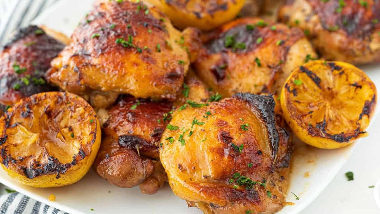 Roast chicken thighs garnished with herbs, served with charred lemon halves on a white plate.