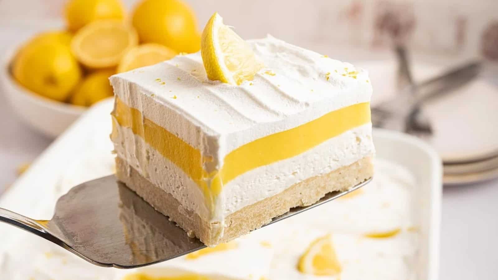 A lemon dessert bar with creamy layers and a lemon wedge on top, held on a spatula over a dish.