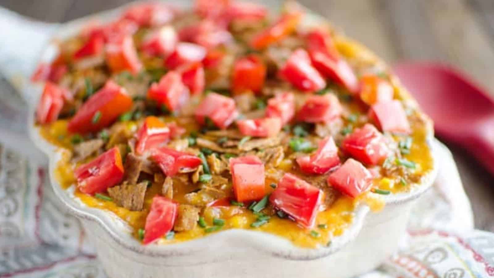 A casserole topped with melted cheese, chopped tomatoes, herbs, and crispy tortilla strips in a ceramic dish.