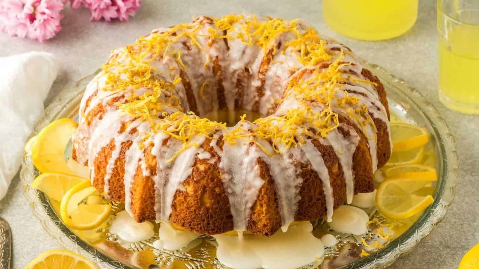 A lemon bundt cake with white glaze, lemon zest, and lemon slices on a glass plate.