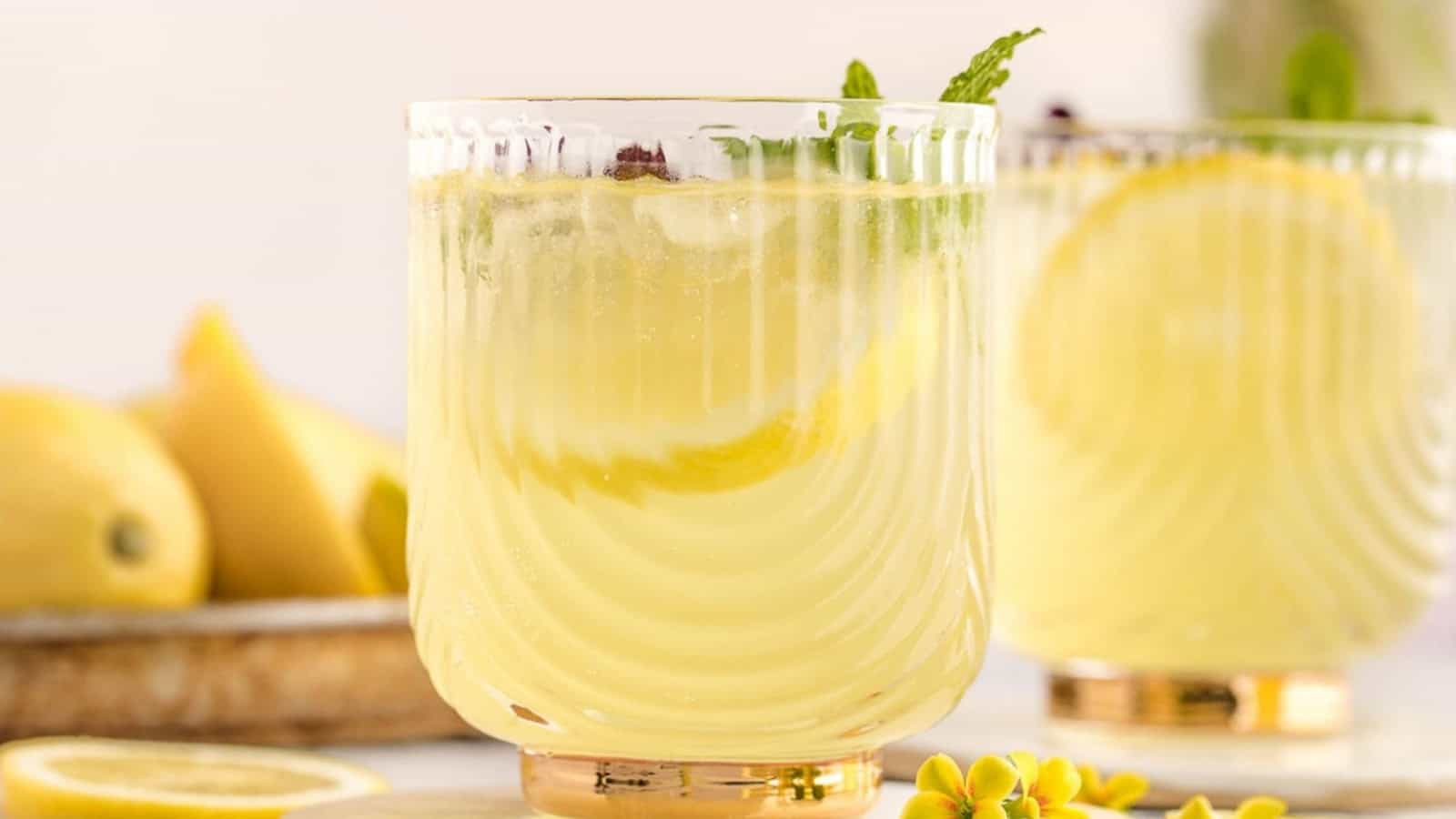 Two glasses of lemonade with lemon slices and mint leaves, with lemons in the background on a light surface.