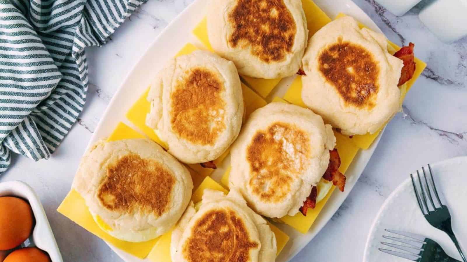 Six breakfast sandwiches with toasted English muffins, eggs, cheese, and bacon on a white platter.