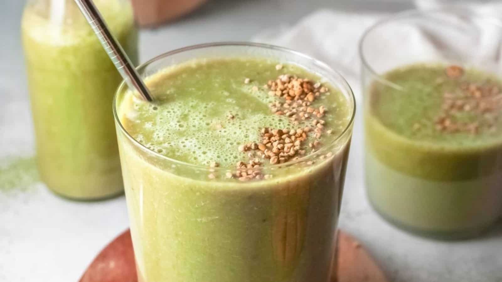 A green smoothie in a glass with a metal straw, topped with seeds, with two more glasses in the background.
