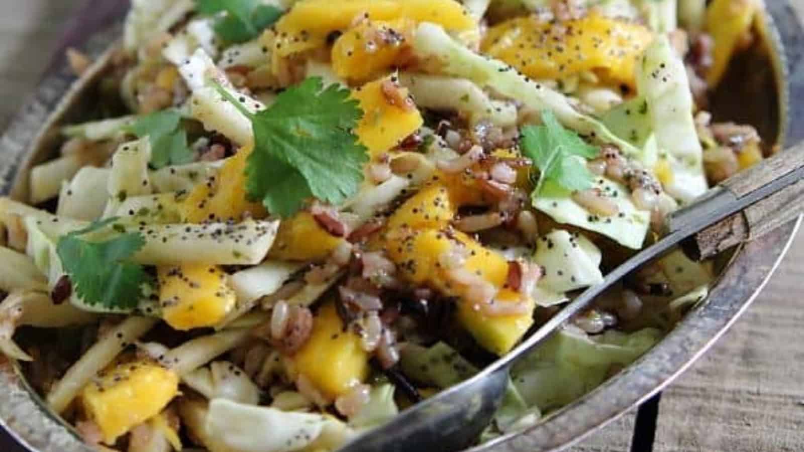 Salad with chopped cabbage, mango, cilantro, wild rice, and poppy seeds in a bowl with a serving spoon.