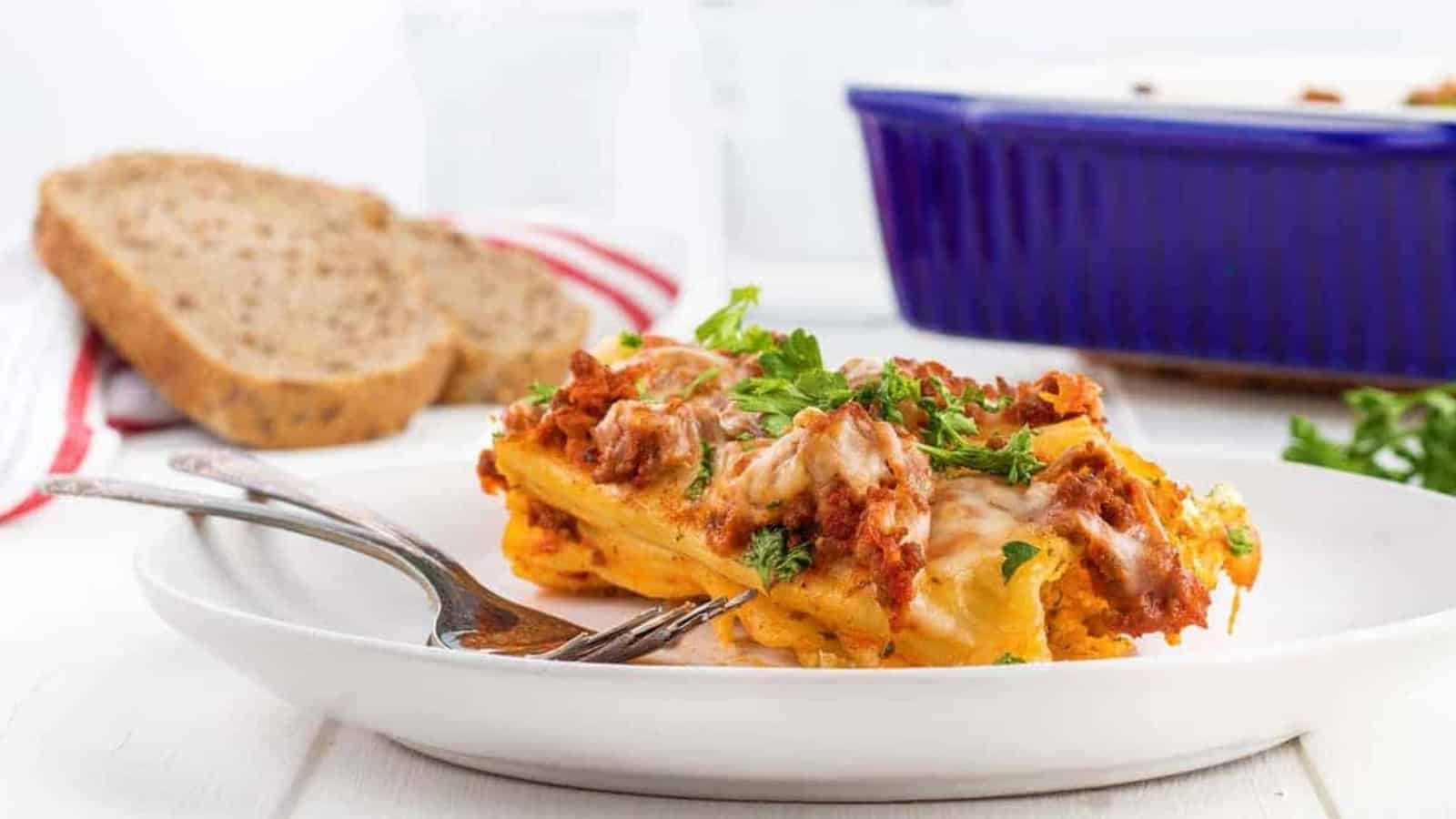 Two pieces of cheesy lasagna on a plate with a fork, with bread slices and a blue baking dish in the background.