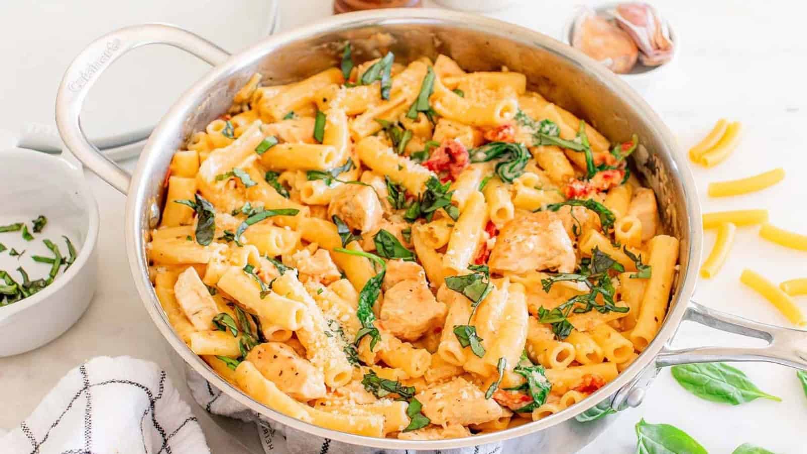 A skillet filled with creamy pasta, chicken pieces, spinach, and sun-dried tomatoes, topped with fresh basil.