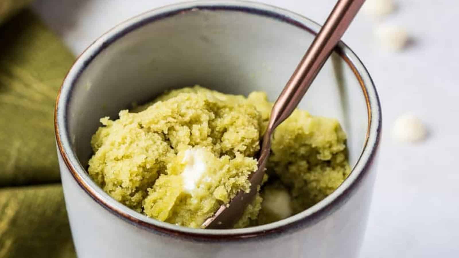 A spoon in a mug with green cookie dough and white chocolate chunks.