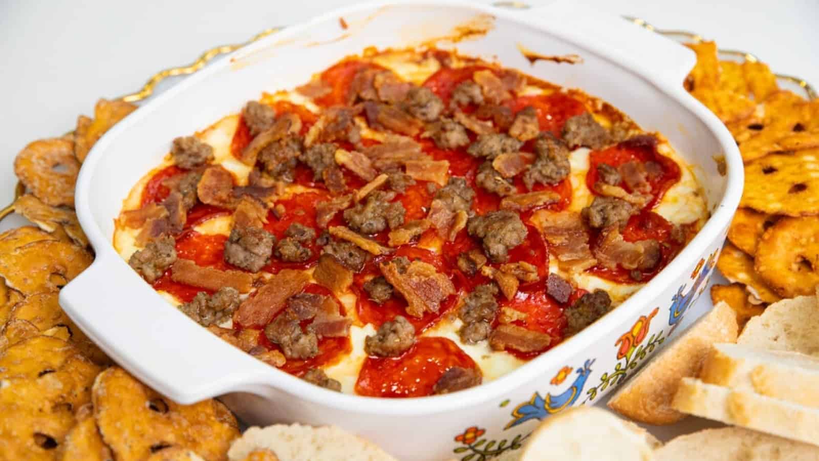 A casserole dish filled with pizza dip, topped with pepperoni, sausage, and bacon, surrounded by bread and crackers.