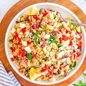 A bowl of quinoa salad with veggies, feta, lemon wedges, and a fork on a wooden board.