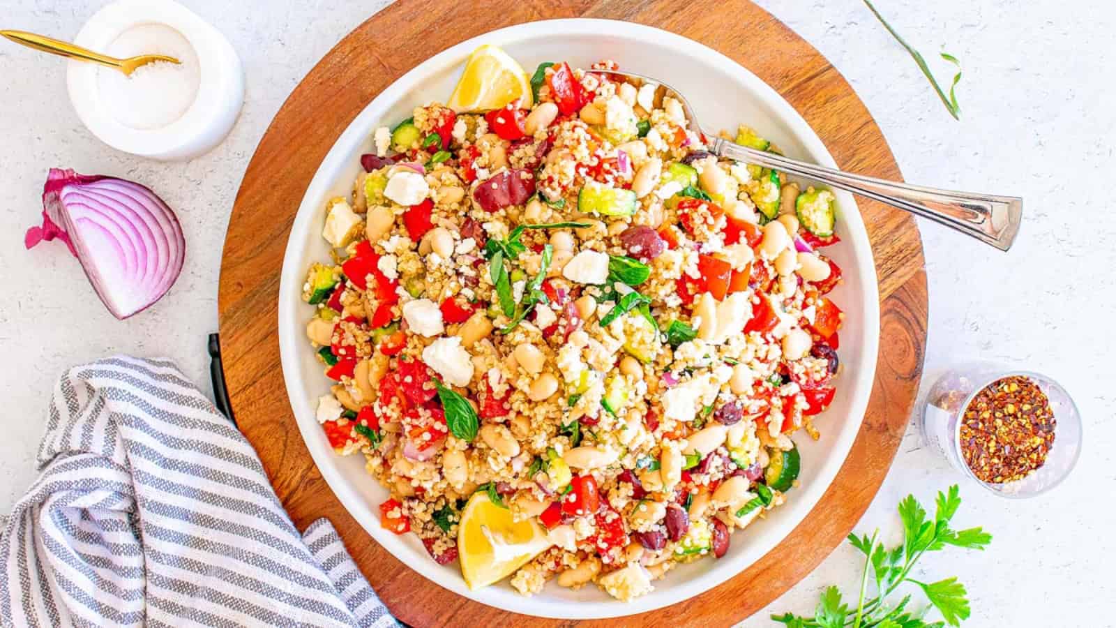 A bowl of quinoa salad with veggies, feta, lemon wedges, and a fork on a wooden board.
