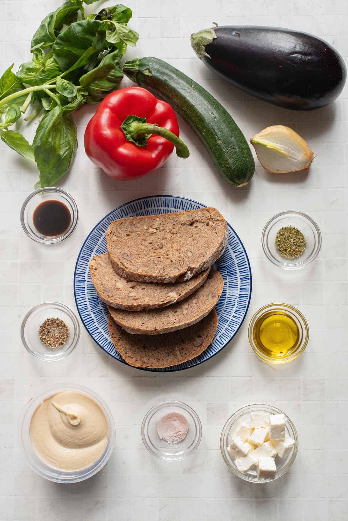 Ingredients for a Veggie Hummus Sandwich, including bread, vegetables, herbs, oil, feta, and seasonings arranged on a white surface.