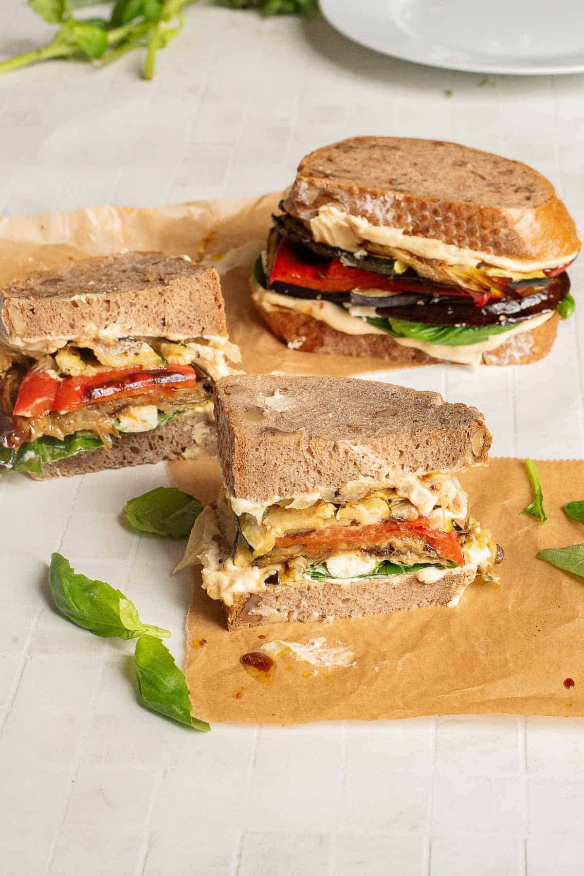 Two Veggie Hummus Sandwiches with grilled vegetables and leafy greens rest on brown paper, with fresh basil leaves scattered around.