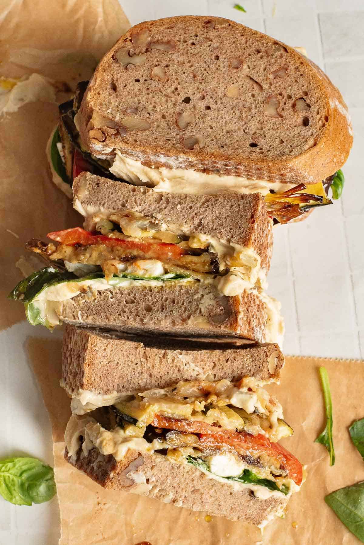 Sliced Veggie Hummus Sandwich with whole grain bread, vegetables, cheese, and greens on parchment paper.