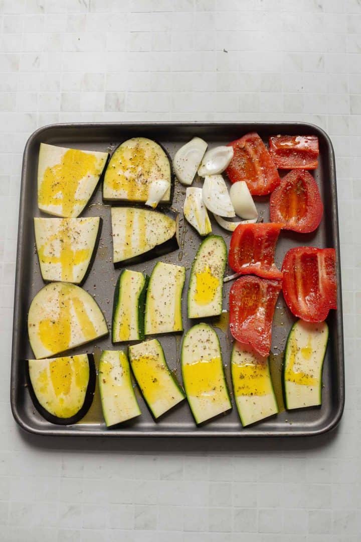 Baking tray with sliced eggplant, zucchini, red bell pepper, and garlic, drizzled with oil and seasoning&mdash;perfect roasted veggies for a delicious Veggie Hummus Sandwich.