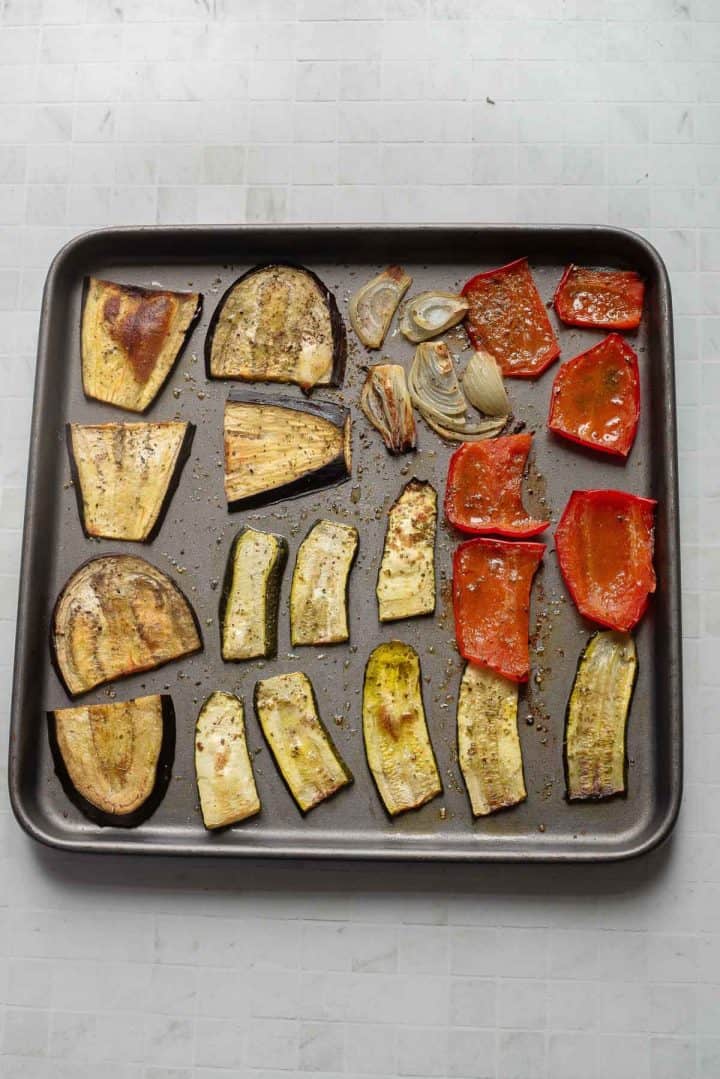 Baking tray with roasted eggplant, zucchini, red bell pepper, and onion slices&mdash;perfect for building a flavorful Veggie Hummus Sandwich&mdash;on a tiled surface.