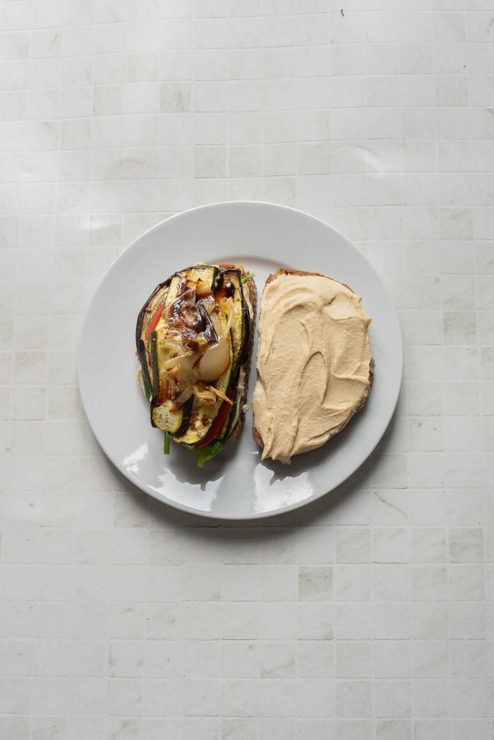 Open Veggie Hummus Sandwich with grilled vegetables and onions on one half, creamy hummus spread on the other, served on a white plate.