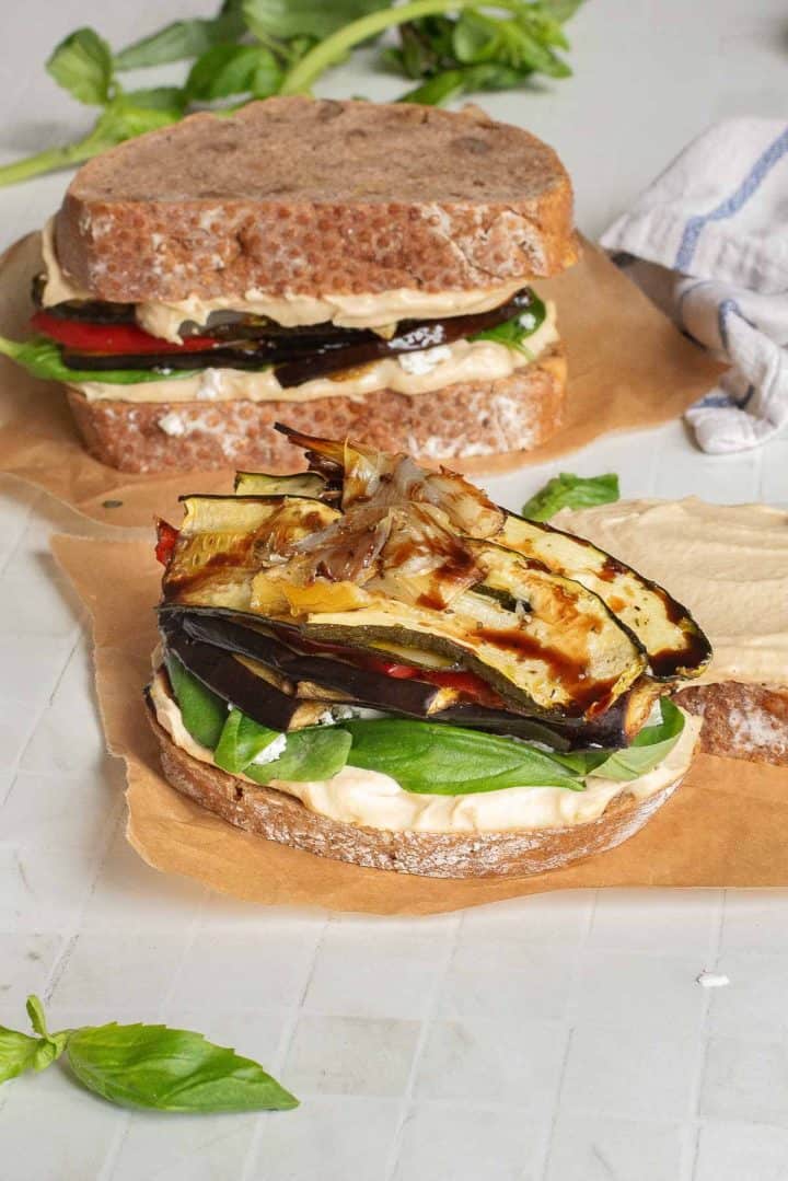 A Veggie Hummus Sandwich with spinach and roasted vegetables on whole grain bread sits in the foreground, with another delicious sandwich in the background.