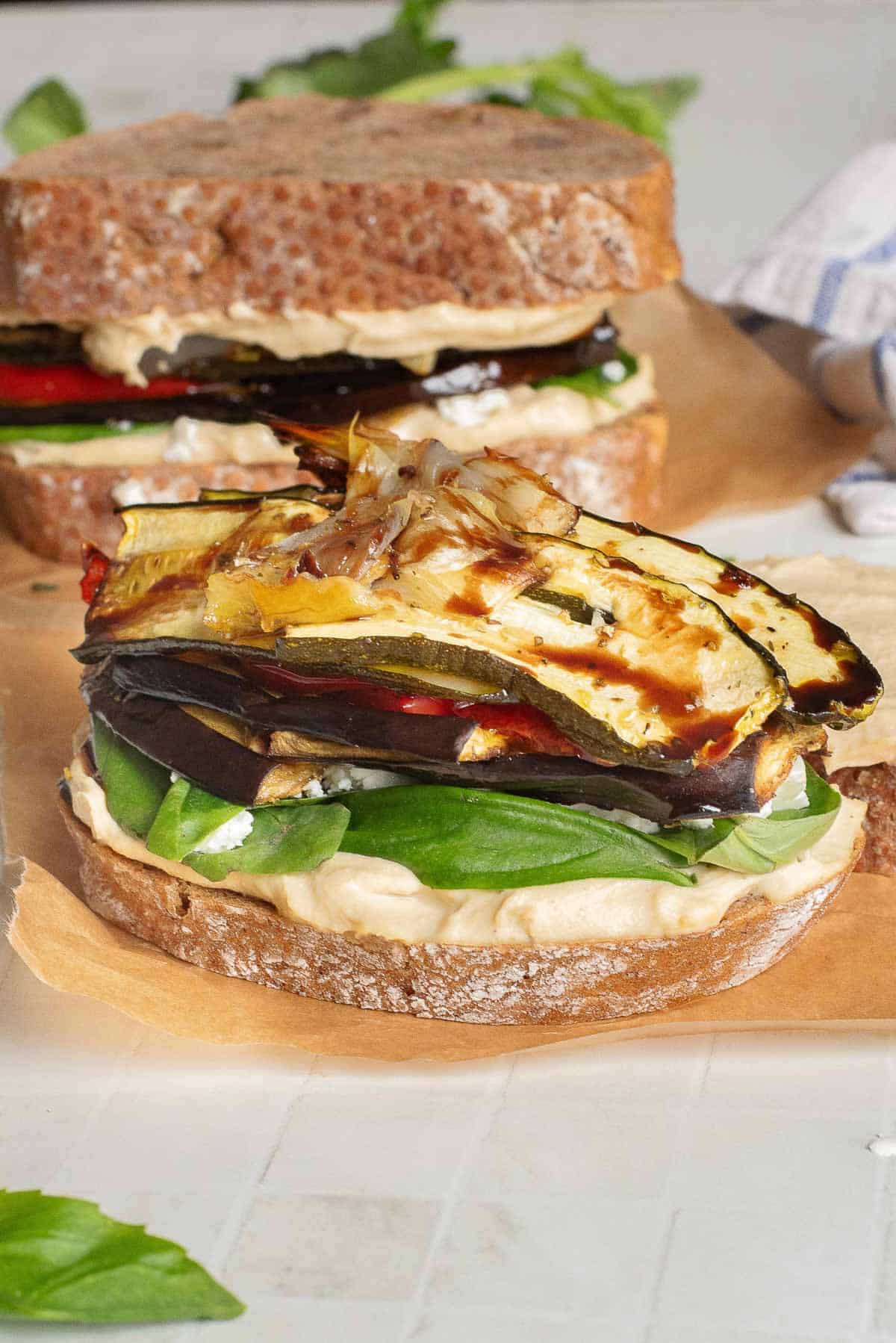 Two Veggie Hummus Sandwiches with spinach and grilled vegetables on whole grain bread, served on parchment paper.