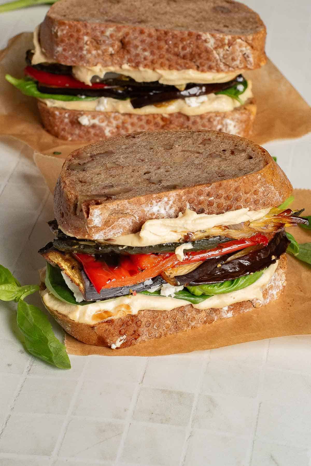 Two stacked Veggie Hummus Sandwiches with grilled vegetables, leafy greens, and spread on rustic bread, set on brown parchment paper.