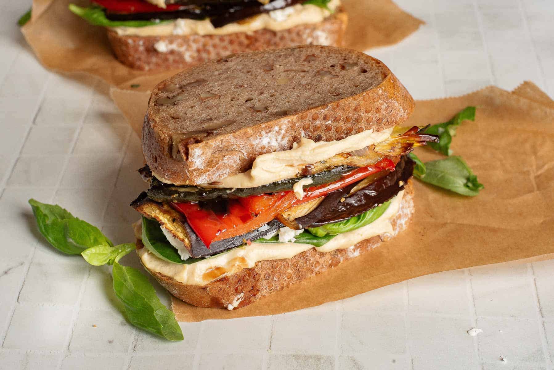 A Veggie Hummus Sandwich with roasted vegetables, leafy greens, and creamy spread on rustic bread, placed on brown parchment paper.