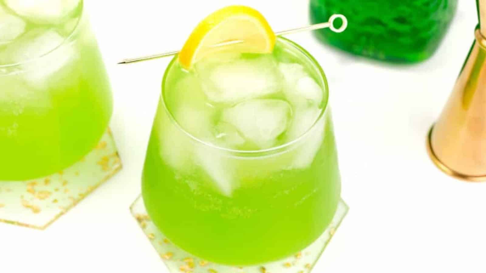 A bright green cocktail with ice, garnished with a lemon slice on a glass, sits on a white and gold coaster.