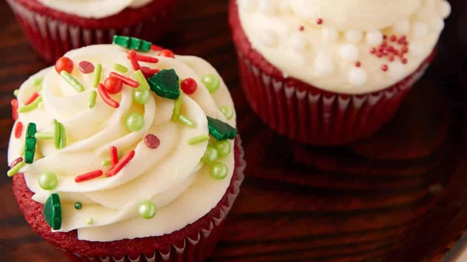 Red velvet cupcakes with swirled white frosting and colorful holiday sprinkles on a wooden surface.