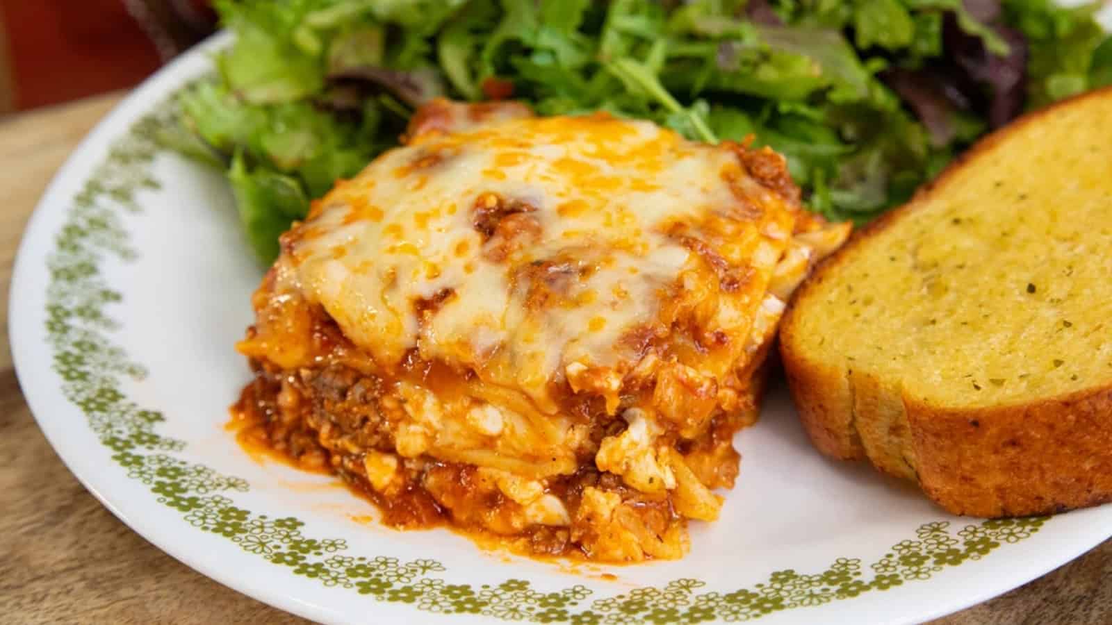 A slice of lasagna with melted cheese, served with salad and a piece of garlic bread on a white plate.
